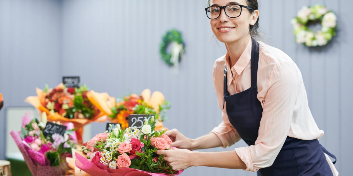 Side view portrait of smiling young woman looking at camera while arranging bouquets in small flower shop, copy space